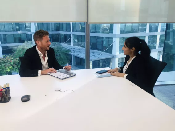 A man and a woman both wearing white shirts and black suit jackets sitting at a large white office table talking to each other.