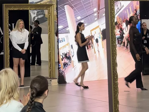 A blonde woman in a white shirt and black skirt receiving applause on a fashion runway