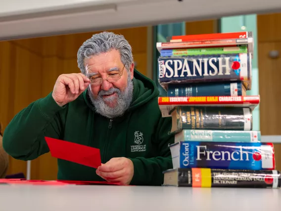 A man with grey hair and a beard sitting at a table looking at a red envelope with a stack of language books beside him.