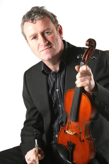 Man - John Carey - wearing a suit holds a fiddle. 