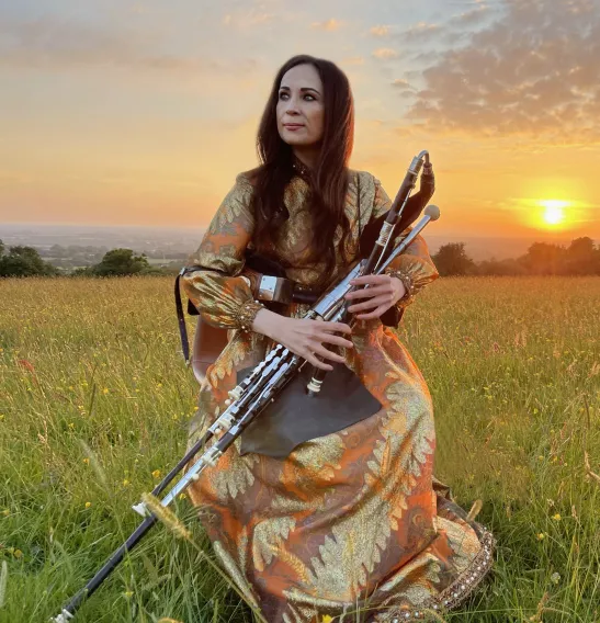 Louise Mulcahy wears a long dress and plays the pipes in a green field