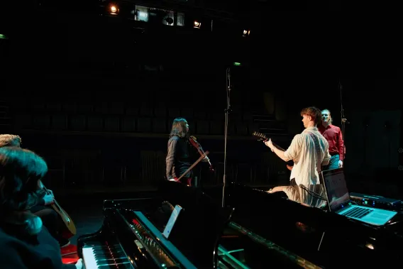 Three performers and musicians on stage while two others sitting next to a piano