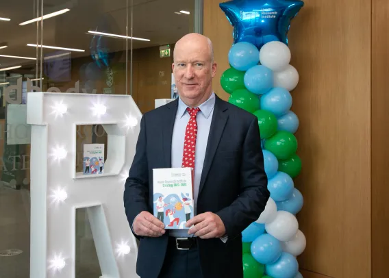 Professor Alan Donnelly posing with the HRI Annual Report in front of a stack of balloons