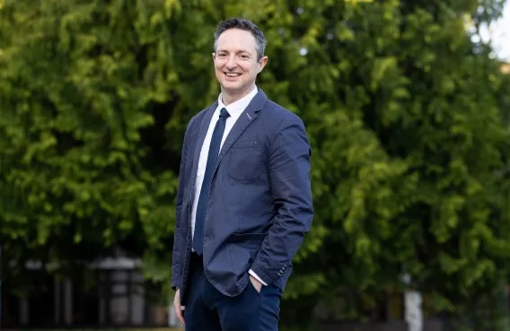 Man with left hand in pocket standing in front of trees and shrubbery