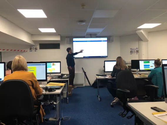 A picture looking up the centre aisle of a classroom. A man is at the front, pointing at slide projected on a screen.