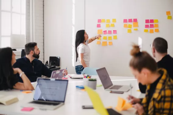 Woman placing sticky notes on a wall as a team of people sitting at a desk with laptops watch her.