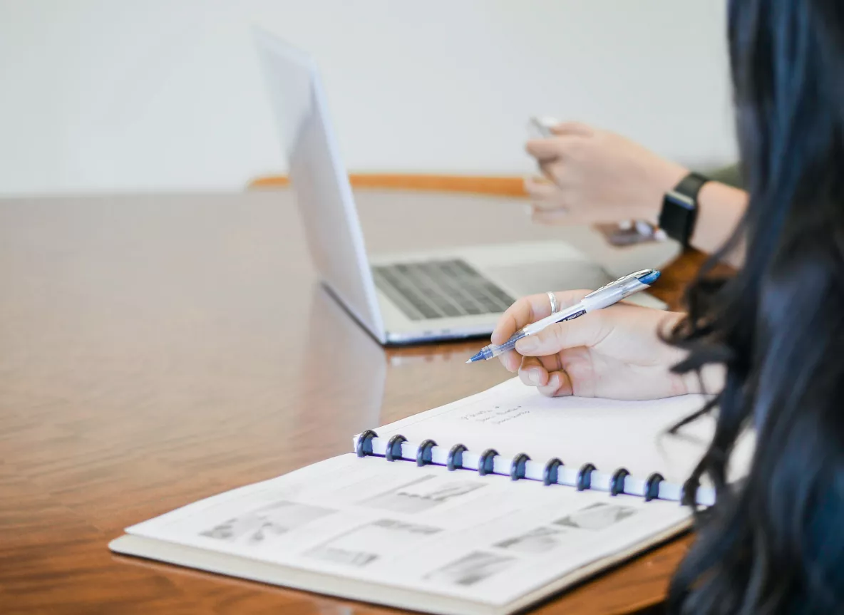 person writing in notebook with another person in background using a laptop