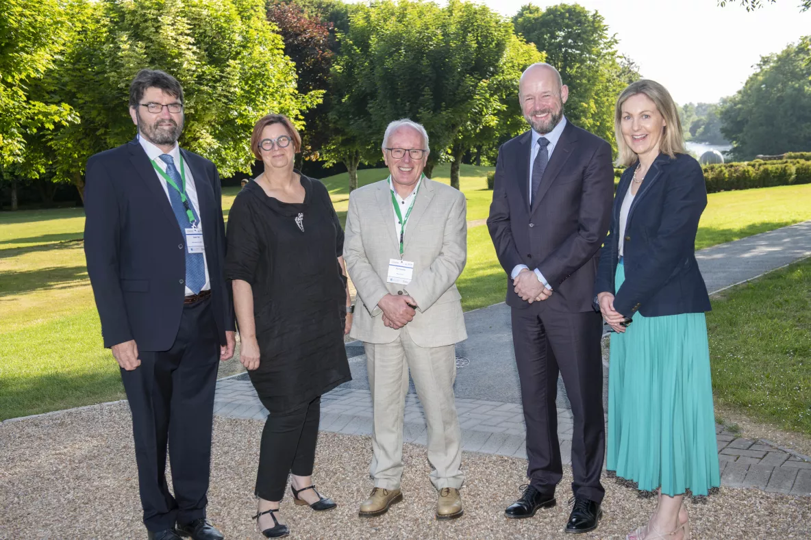 Photos of five individuals standing together with Green lawn and bushes in the background. 