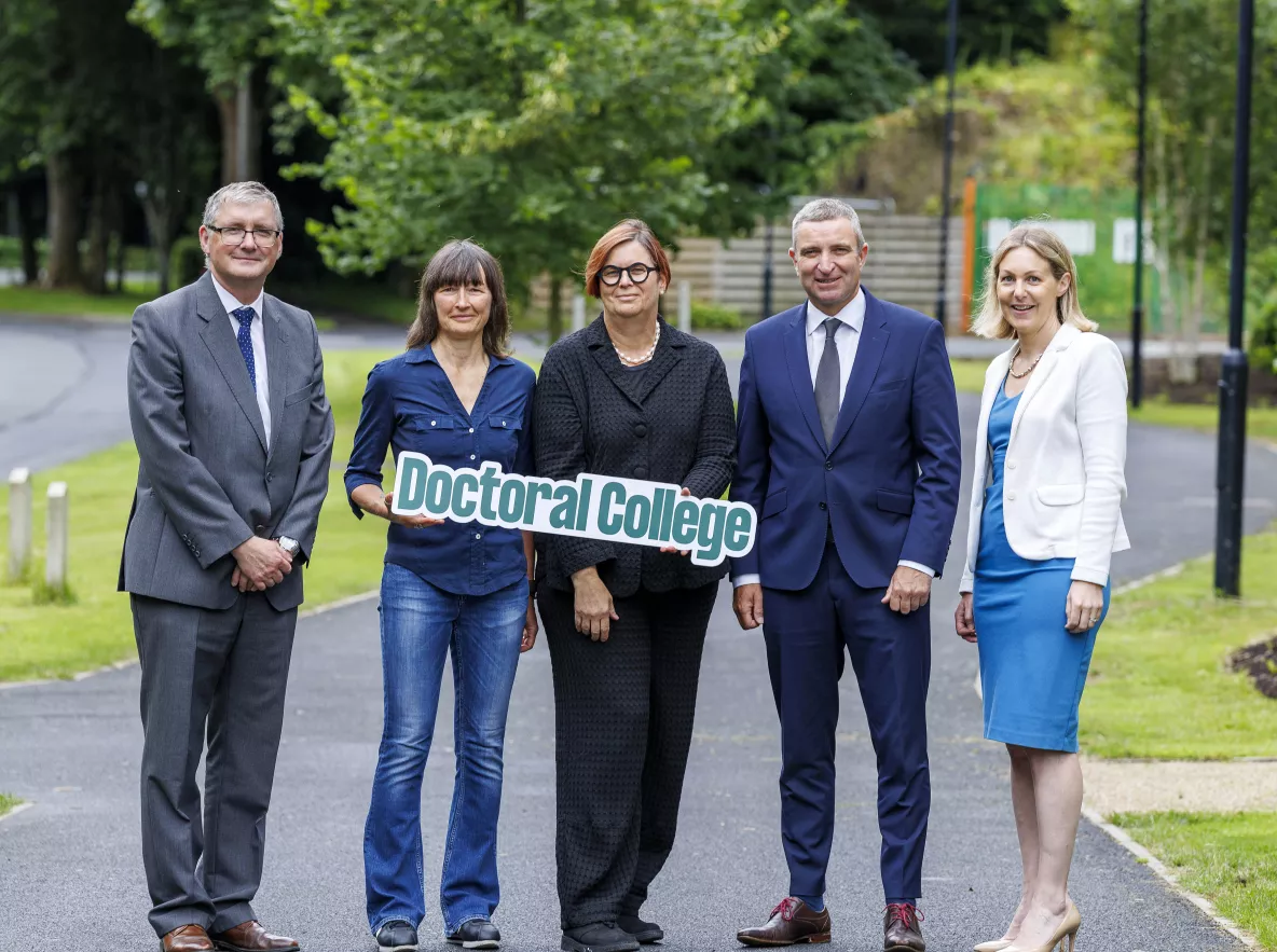 The subject of the photo are standing in a line together smiling towards the camera holding a sign that says Doctoral College. 