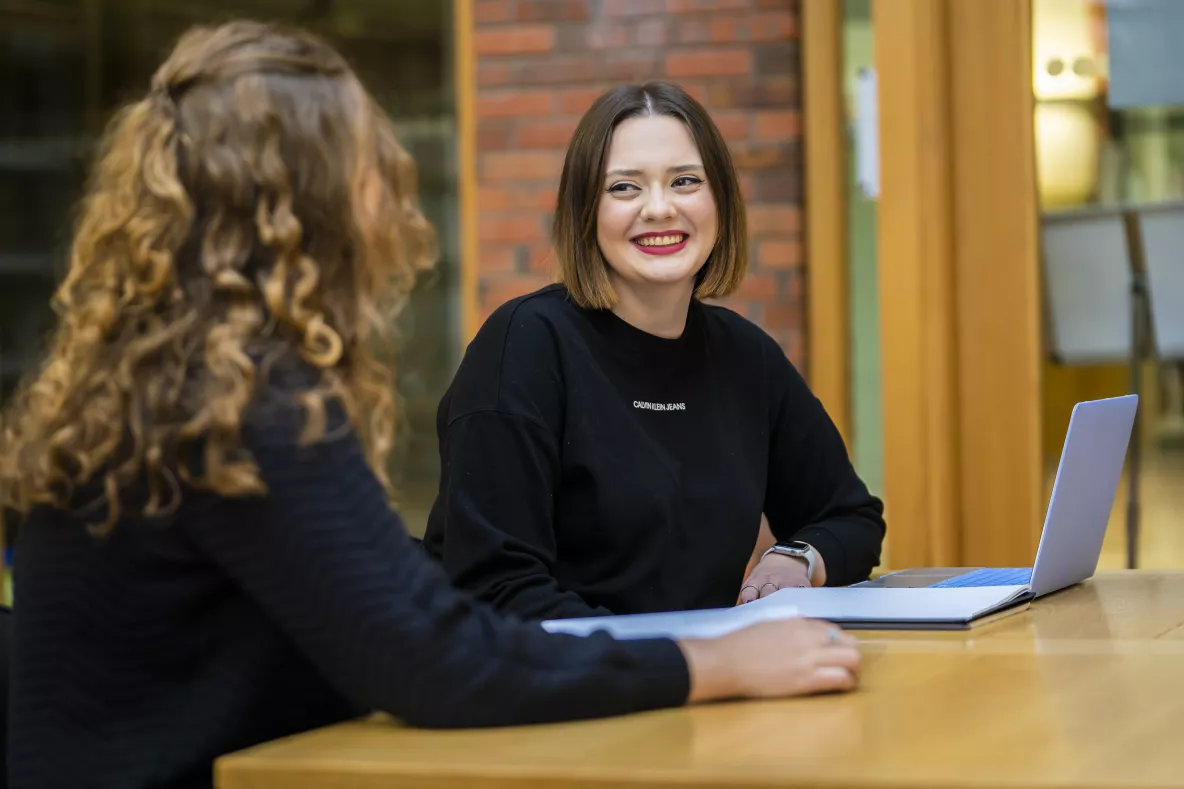 2 students chatting in library in front of laptop