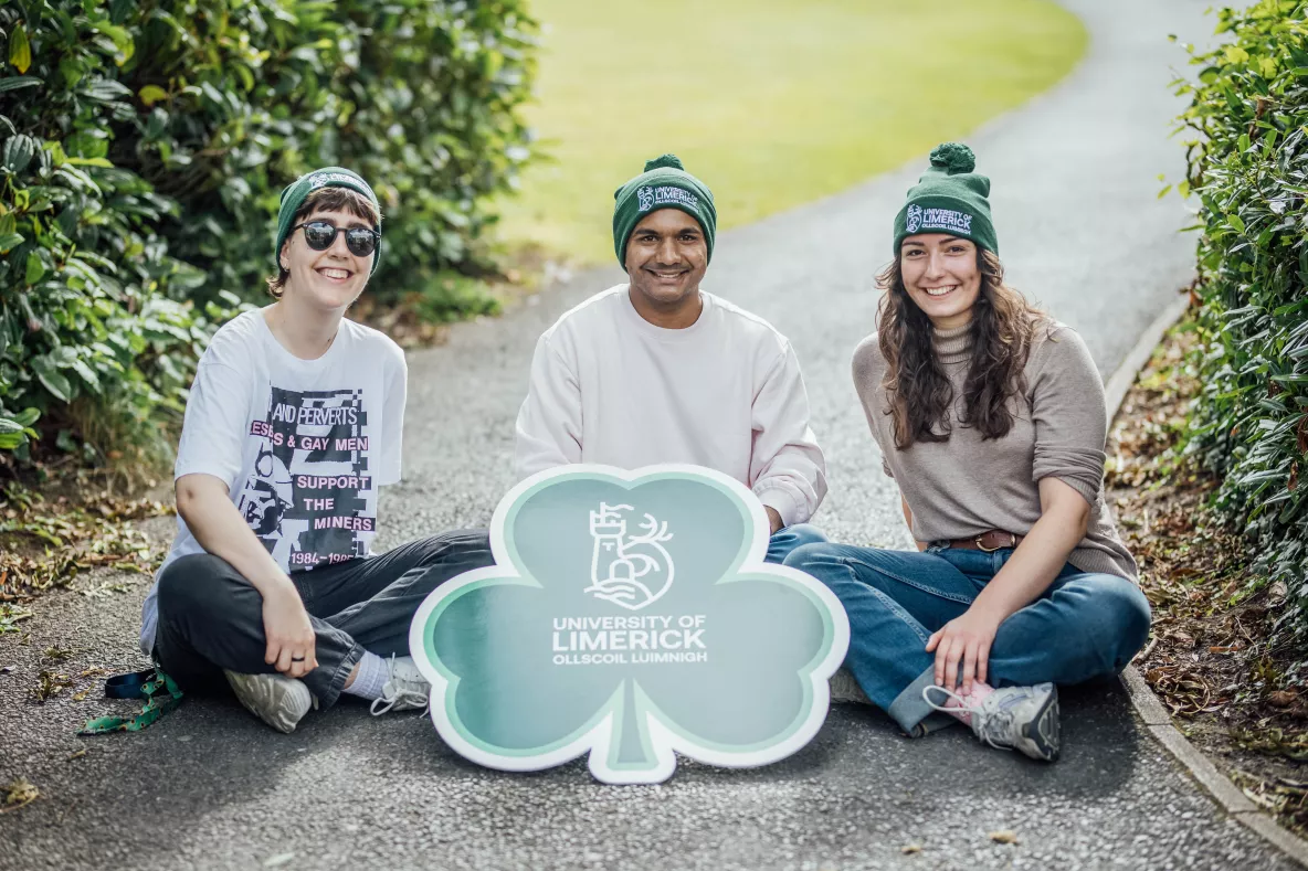 3 students wearing UL green beanies sitting on the ground with a shamrock sign with the UL logo on it