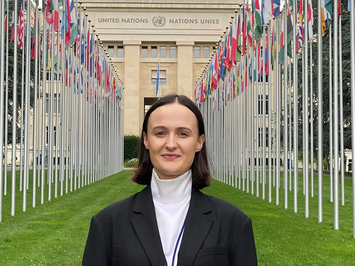 A woman with short brown hair in a black jacket and white top standing in front of the United Nations Building in Geneva