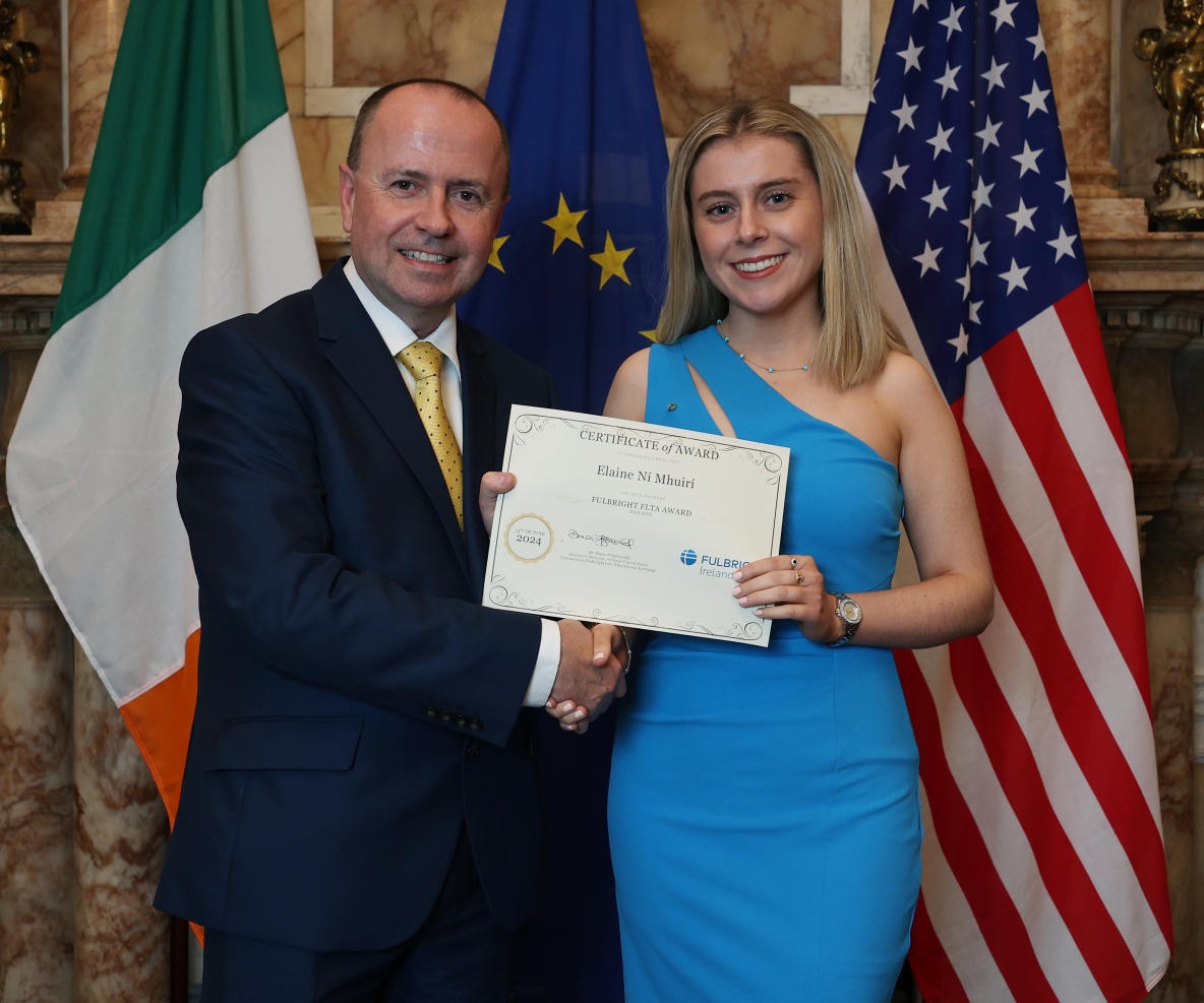 A photo of a man in a black suit, white shirt and gold tie, standing beside a women in a blue dress. They are shaking hands and holding an award certificate between them, in front of the flags of Ireland, the European Union, and the United States. Paul is wearing a black suit and gold tie, hol