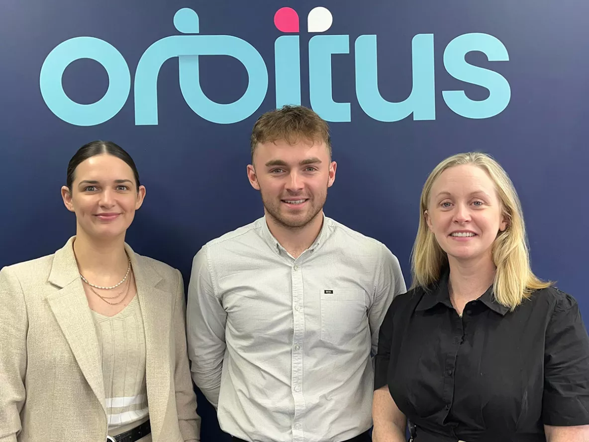 Two women and a man standing side by side in front of a wall with a sign that reads Orbitus