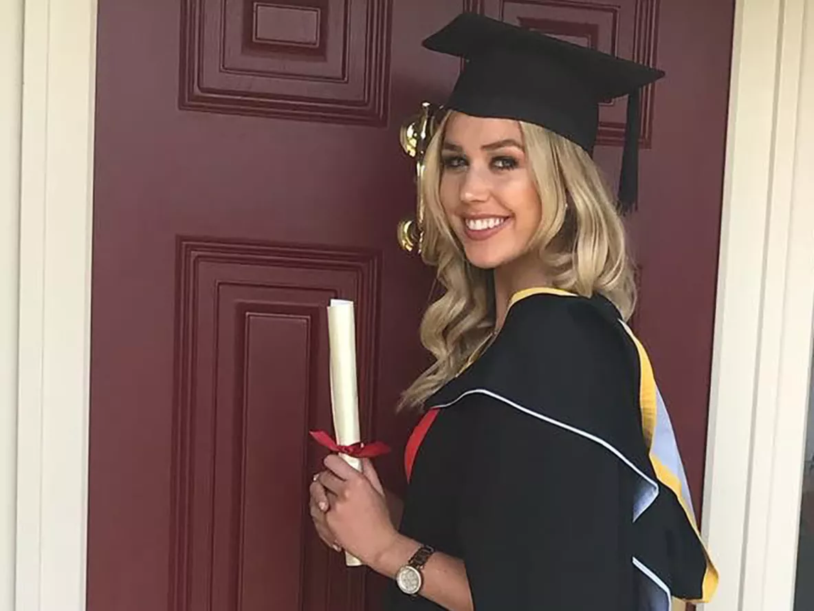 A woman with blonde hair wearing a black graduation cap and gown holding a diploma in front of a red door