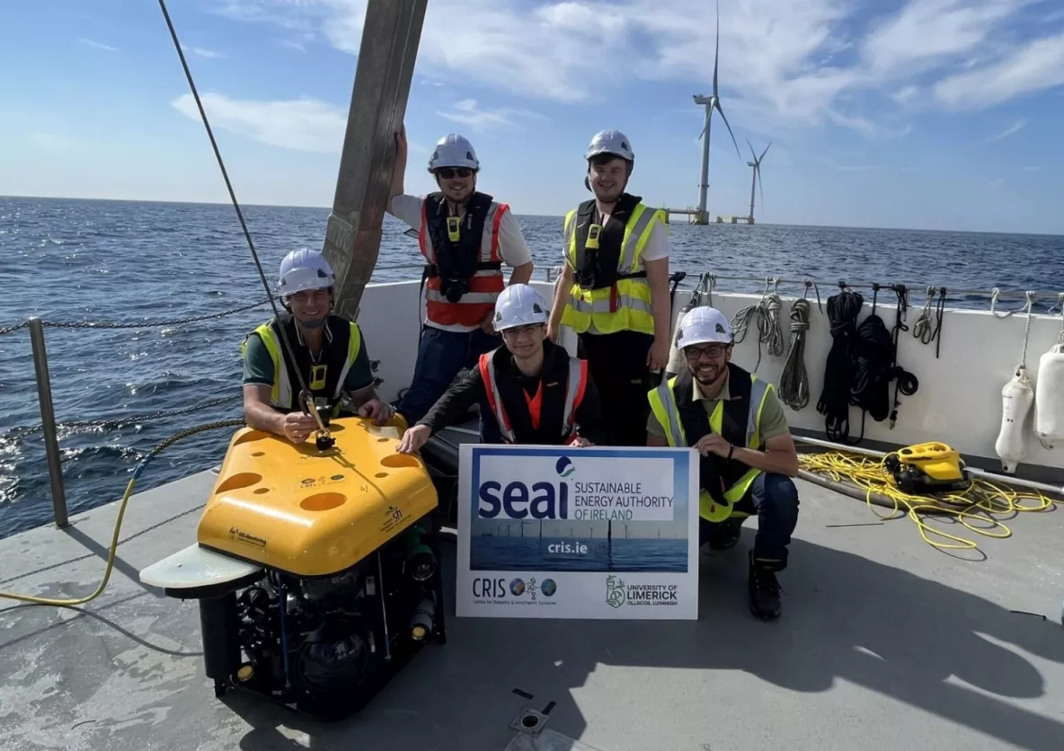 Five people wearing hard hats and hi visibility jackets and life jackets aboard a platform on an off-shore wind farm in the Atlantic Ocean with wind turbines in the background