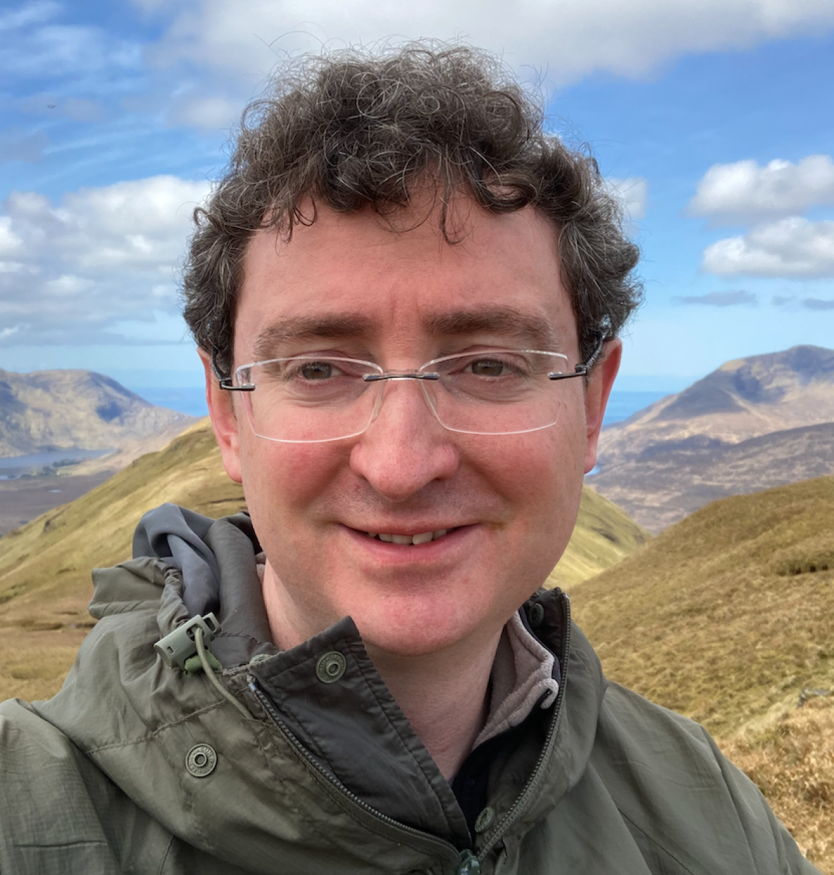 A photo of a man with glasses, wearing a jacket, smiling to the camera against a backdrop of mountains