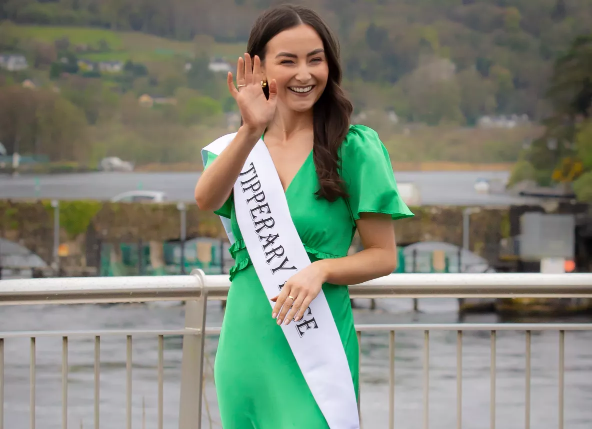 A photo of a person in a green dress and white sash, waving, with trees and a bridge over water in the background 