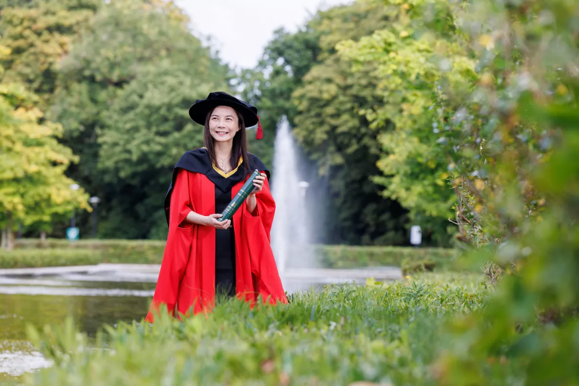 A photo of a woman wearing red graduation robes and black hat, smiling and holding a green scroll. She is standing against a backdrop of a water fountain and green trees and foliage.