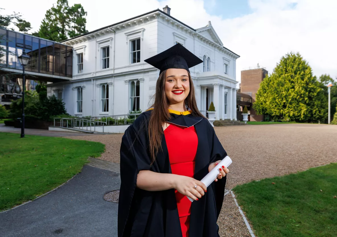 A woman in a red dress wearing a black graduation cap and gown standing in front of a white building.