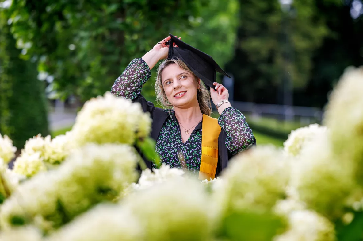 Picture of women wearing graduation robes with flowers out of focus in the foreground and trees in the background