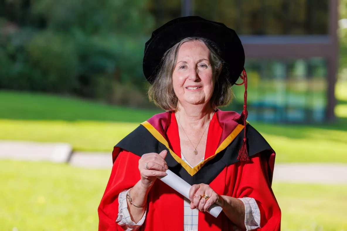 A photo of a woman in red ceremonial robes and a black graduation cap, holding a white scroll and smiling. In the background are grey steps and green grass and trees.