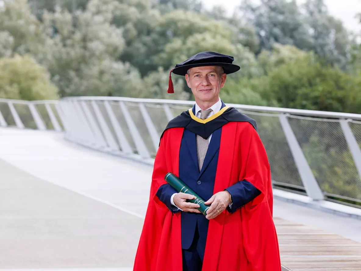 A man in a red graduation gown wearing a navy suit standing on a bridge with trees in the background