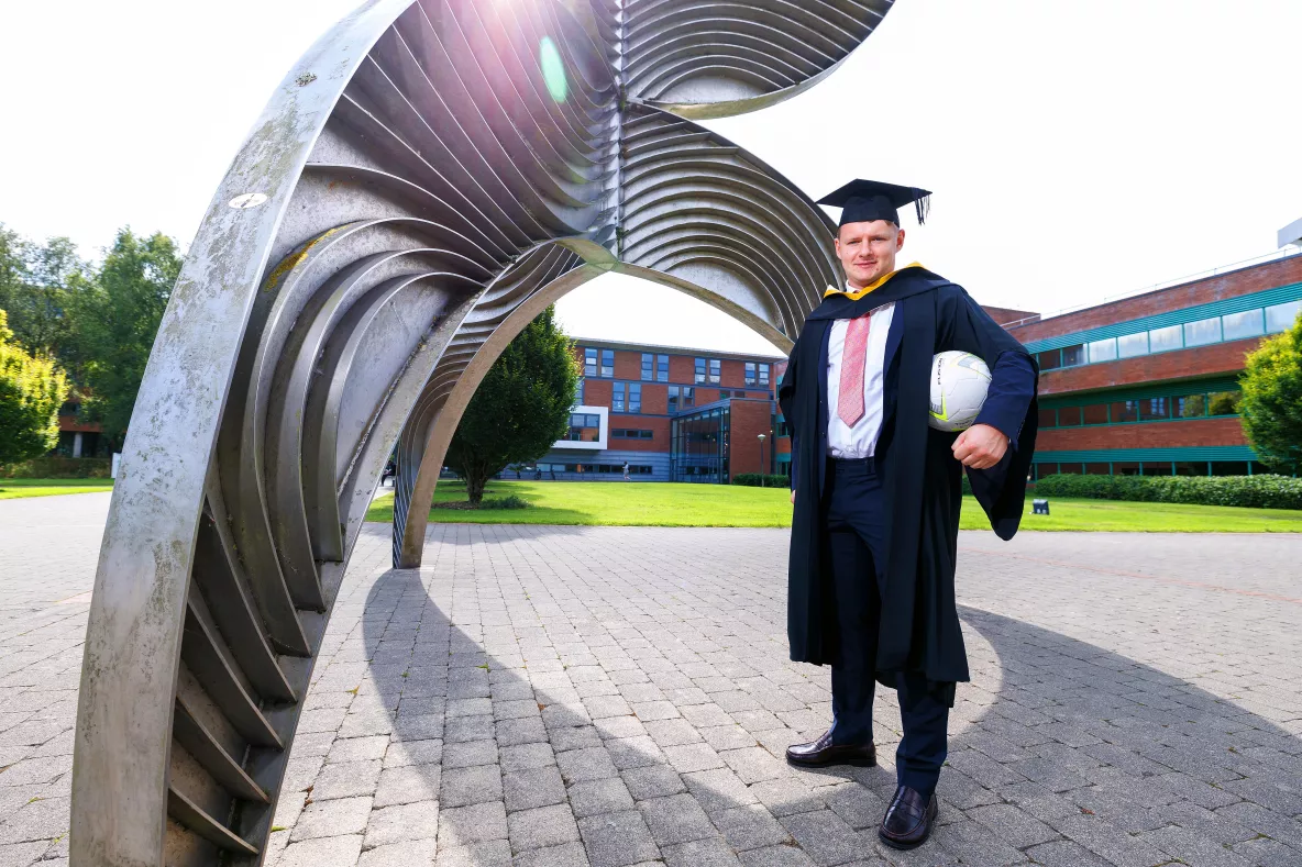 A photo of a man in a black suit, white shirt and pink tie underneath black graduation robes, wearing a graduation cap and dark shoes. He is holding a soccer ball under his left arm, standing in front of a tall metal sculpture. In the background is a red brick building with windows.