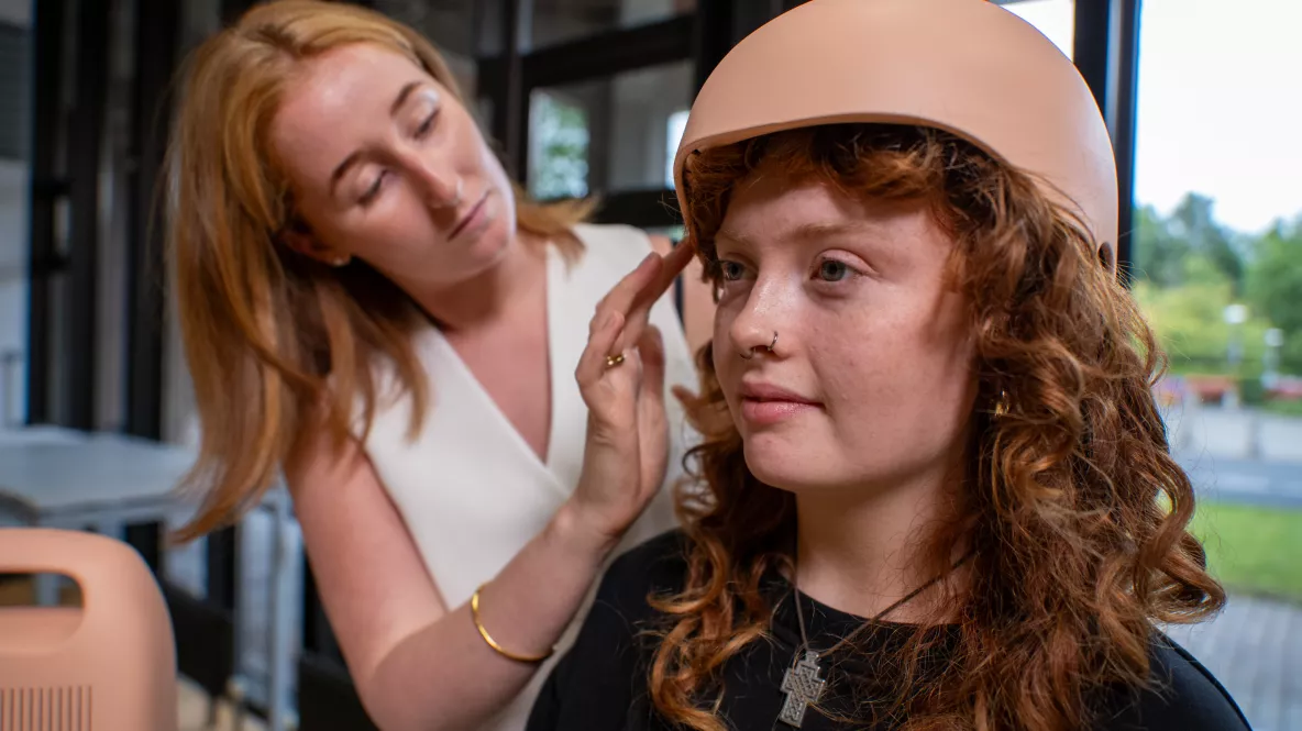 A photograph of two women with red hair. On the right, closest to the camera, is a woman in a black top sat under a pale pink helmet. She is being attended to by a woman in a white top stood behind her, who is tucking her hair into the helmet. The helmet is scalp-cooling device for people undergoing chemotherapy treatment