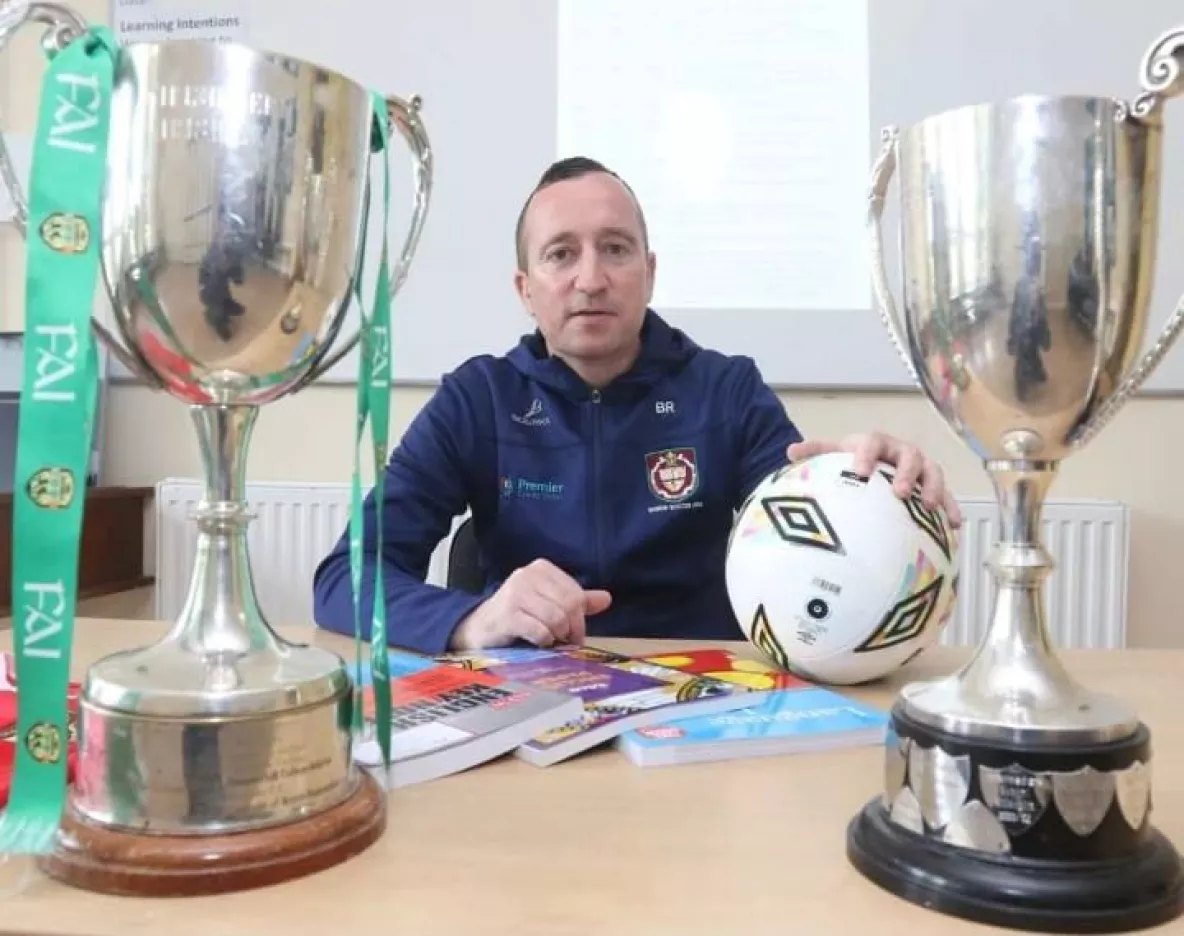 Man sitting behind desk with two football champion cups in front of him