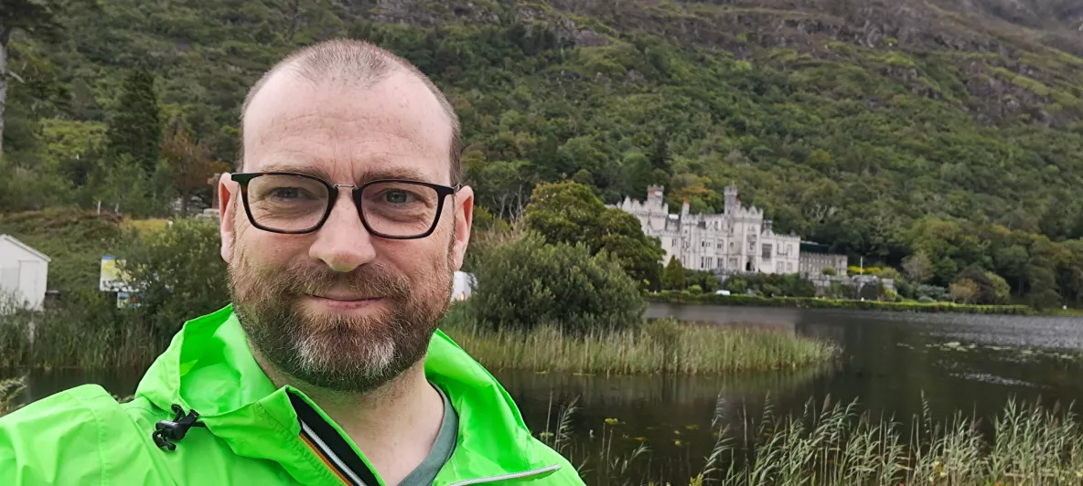 Man wearing glasses with bright green jacket on stands facing camera with an old building behind him in a lakeside location