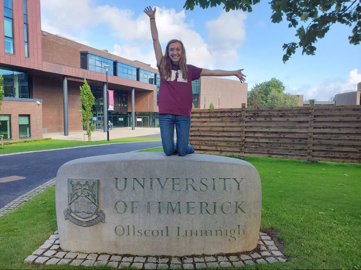 A woman in a wine t-shirt with UL on it kneeling on a University of Limerick sculpture