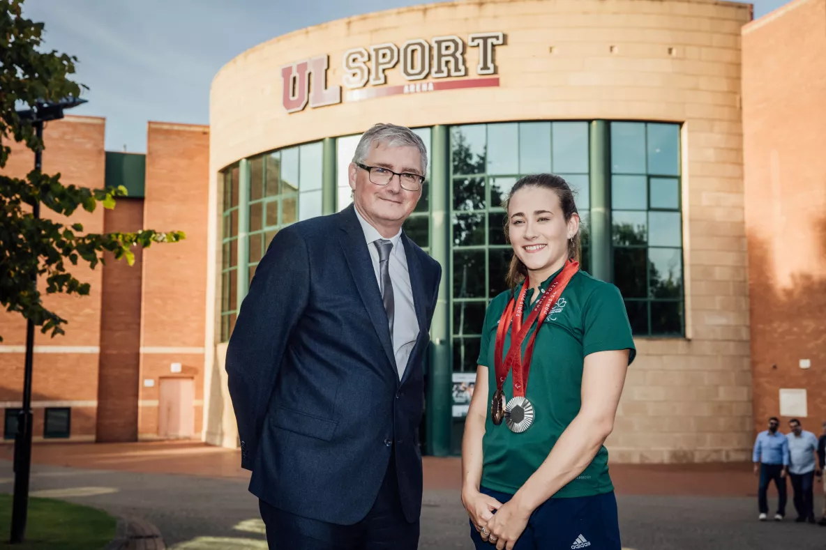 Man with glasses and wearing suit to the left and young girl to the right wearing paralmpic medals around her neck in university sports setting