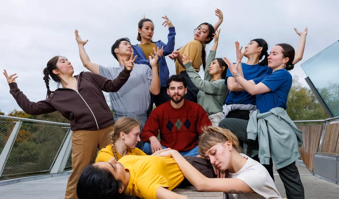 A photo of a man in a red jumper looking directly ahead, surrounded by dancers pulling various dance shapes with their arms. In the background is a bridge and green foliage.