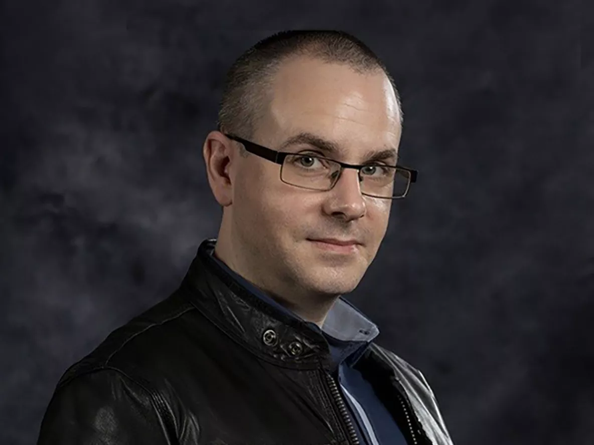 A man wearing glasses and a black leather jacket standing in front of a dark grey backdrop looking at the camera