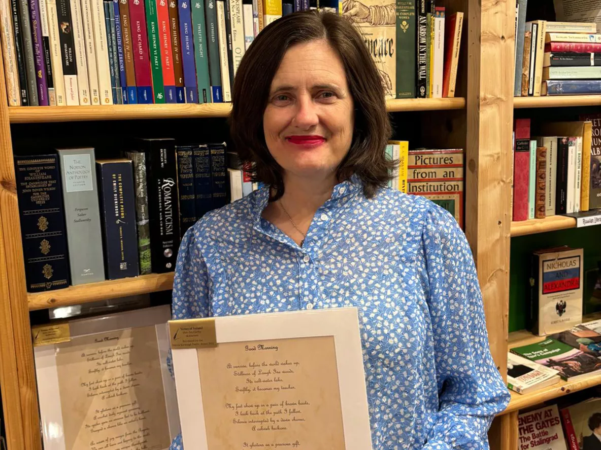 A woman with shoulder length brown hair wearing a floral white and blue shirt standing in front of a bookcase holding a printed poem.