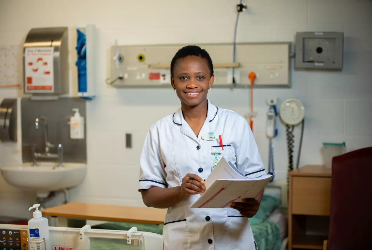 Woman wearing a white nurses uniform standing in a hospital ward holding a pen and chart