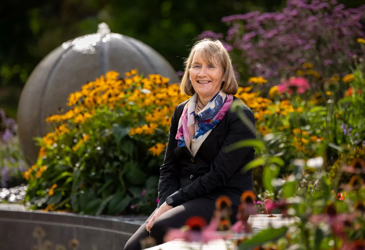 Woman with shoulder length brown hair, wearing a a black jacket and trousers and multi-coloured scarf sitting amongst outside on a sunny day surrounded by flowers and plants