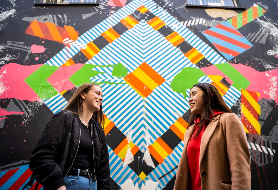 Two girls looking at each other on front of a colourful grafittied wall