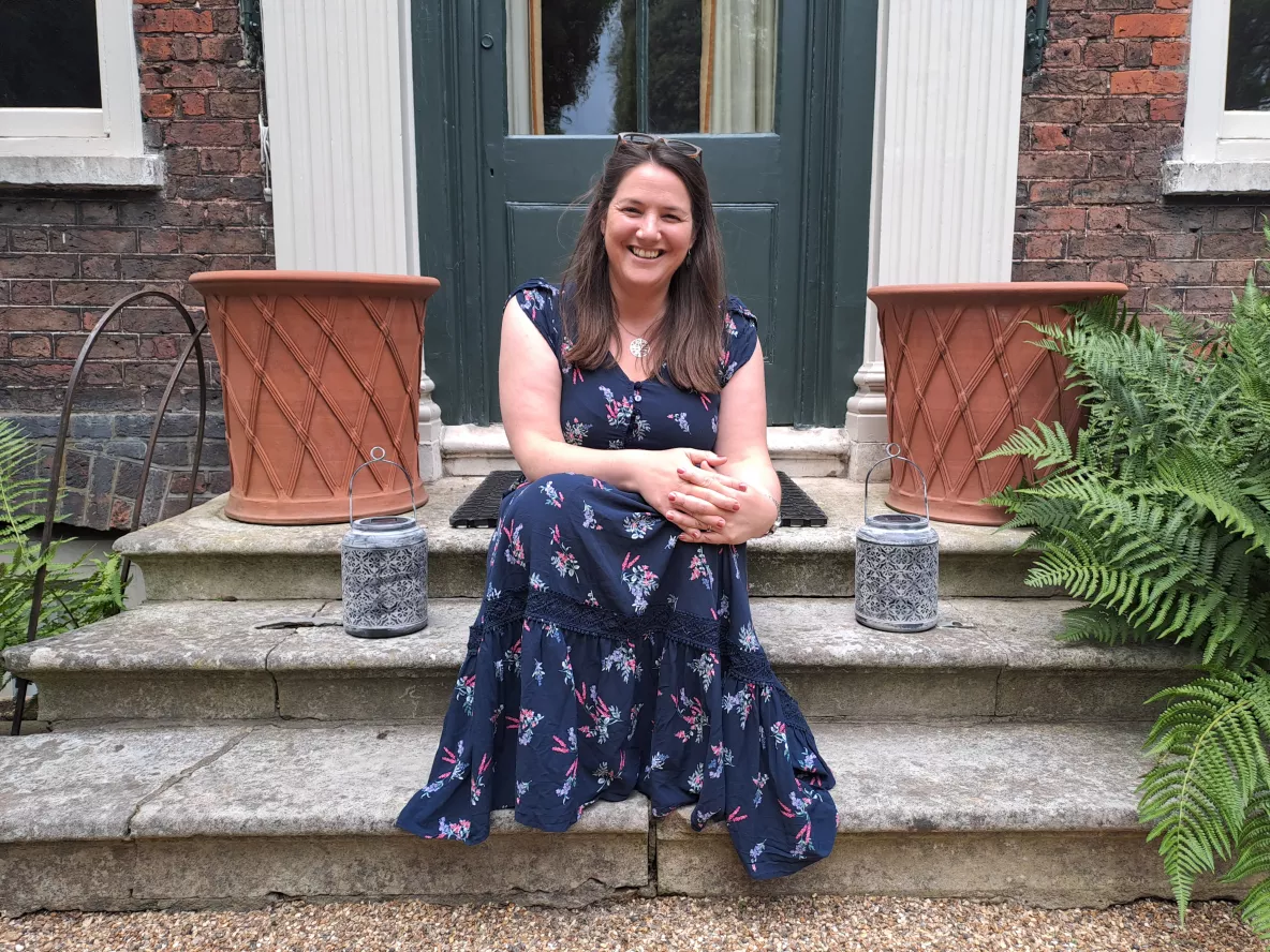 A photo of a woman with long, brown hair wearing a short-sleeved navy floral dress, sitting on a stone step. Behind her is a dark-coloured door and a terracotta pot on either side.