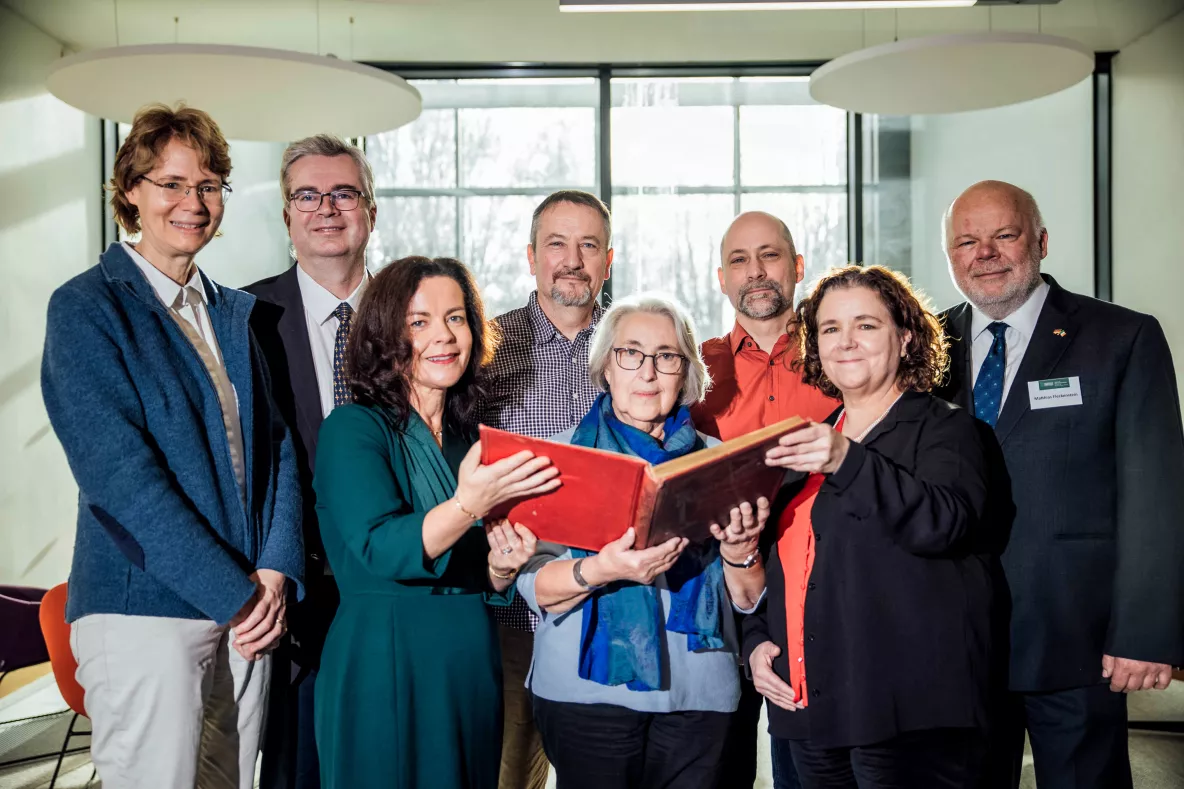 A photo of a group of people - four women and four men - standing in two rows. The three women in the front row are holding up a red book between them