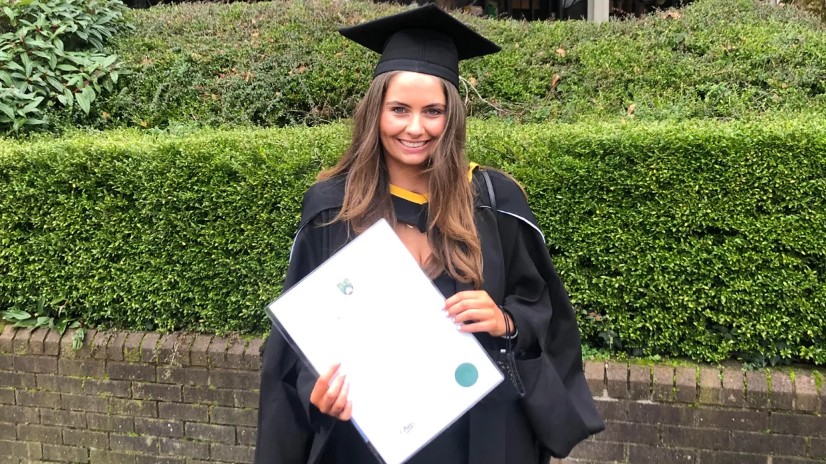 A photo of a female graduate. She is wearing black ceremonial conferring robes, holding her parchment, against a brick wall and green hedge.