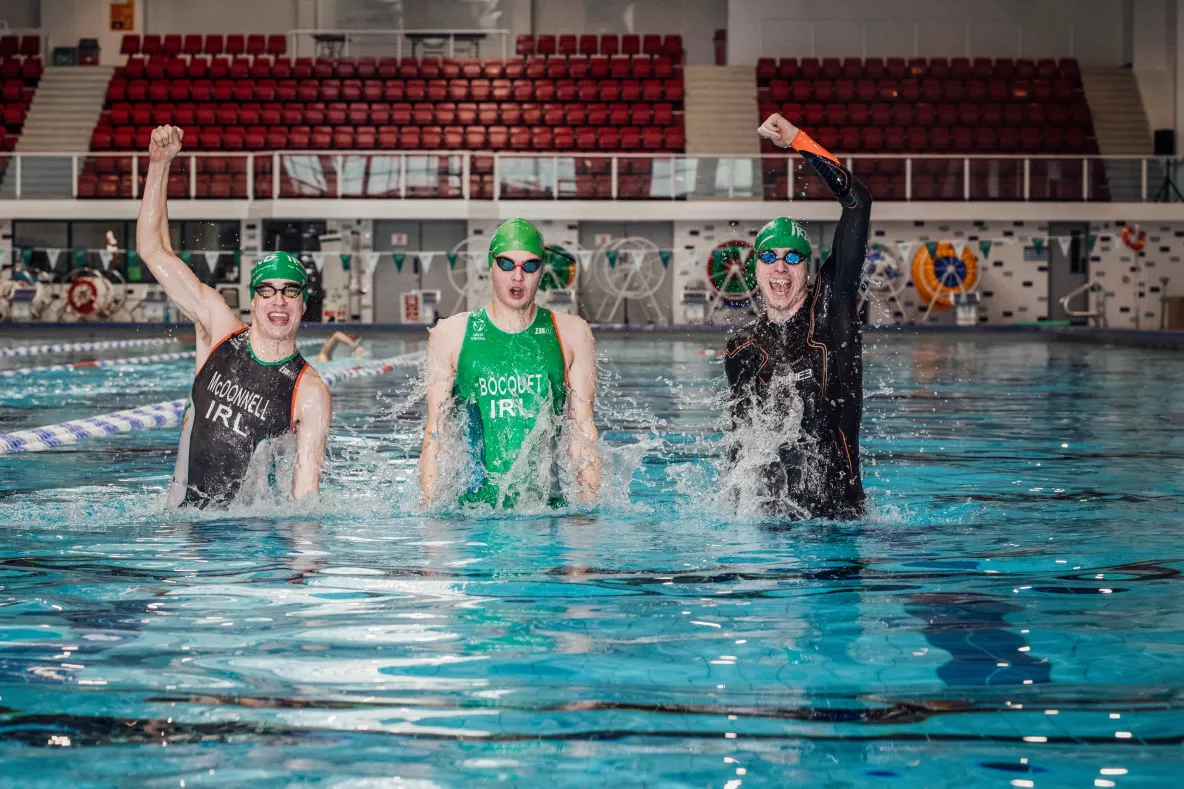 Three male triathletes wearing traithlon suits, goggles and swim hats jumping up in a large swimming pool