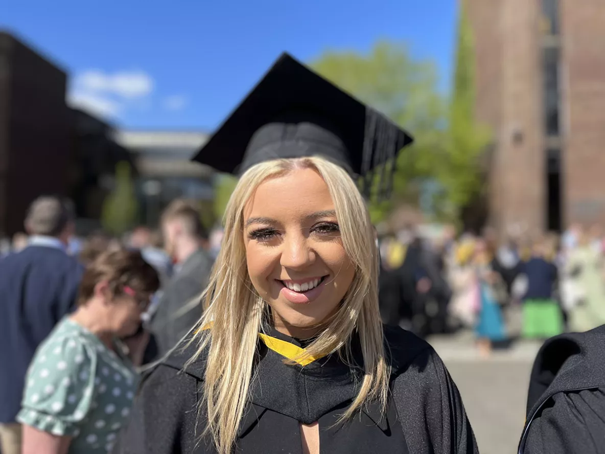 A woman with blonde hair wearing a black graduation cap and gown smiling at the camera