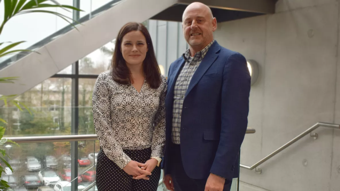 A photo of a woman and a man in smart dress, standing together against a grey wall and window.