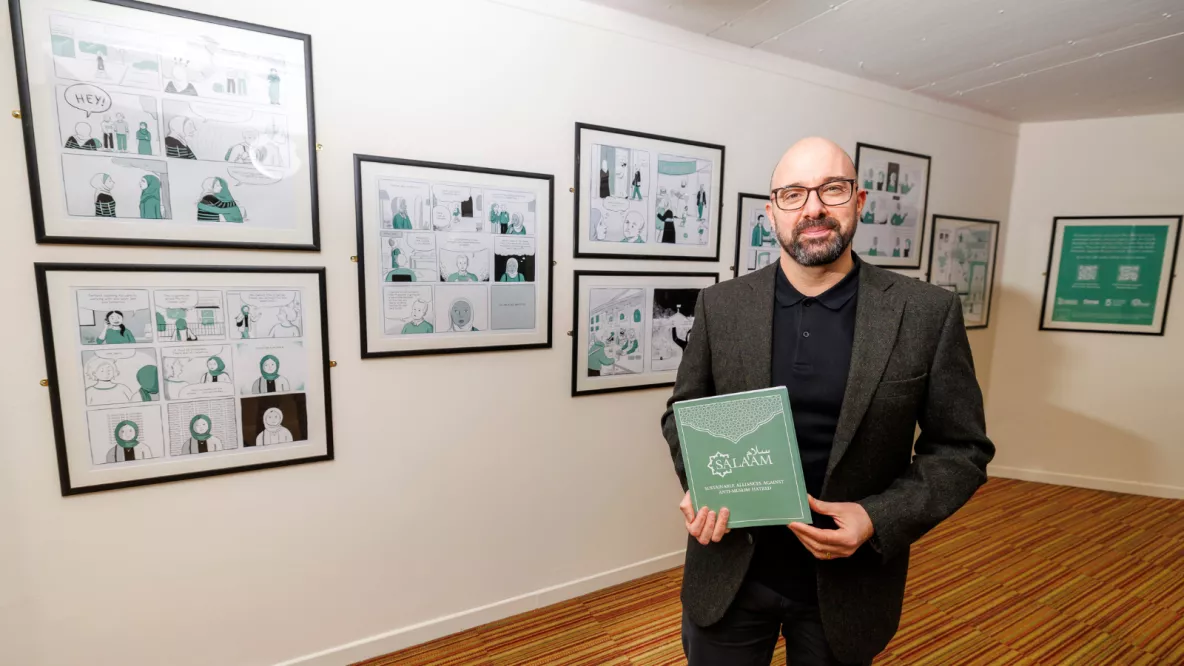 A photo of a man in smart dress, standing in front of a wall of framed pictures, holding a paper booklet.