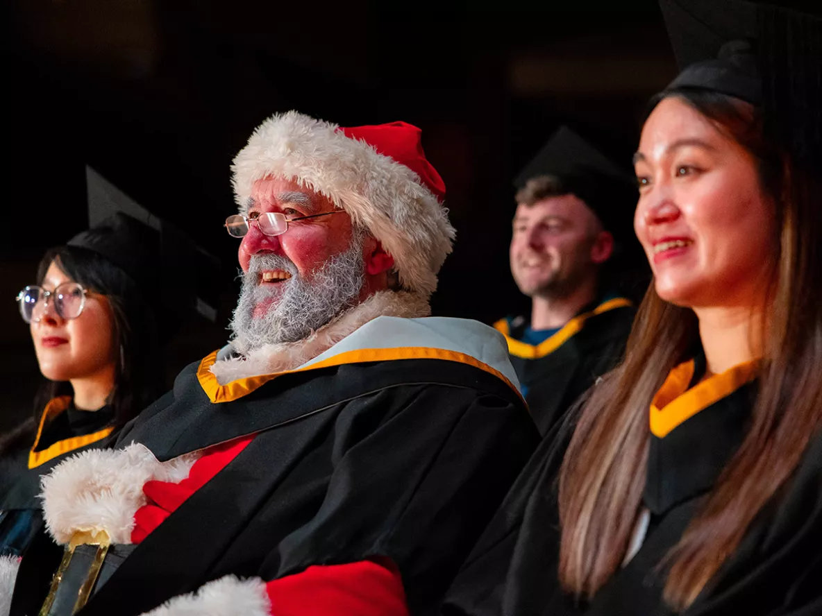 A woman with dark hair and glasses, a man dressed in a Santa suit, and a woman with long brown hair, all sitting in a row looking to the side of the camera wearing graduation gowns. 