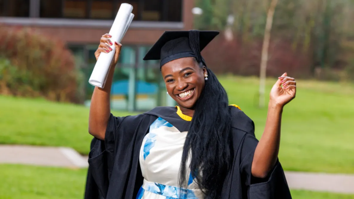 A photo of a women smiling, with her hands in the air, wearing black ceremonial robes and cap over a blue and white dress. She is holding a white scroll in one hand.