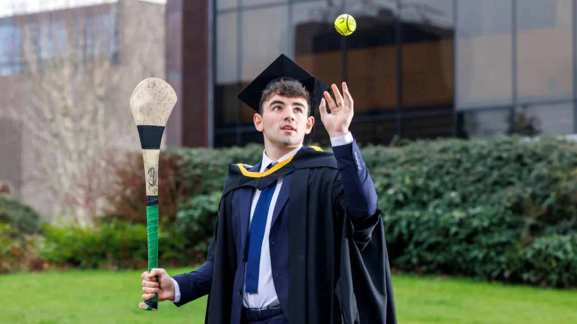 A photo of a man dressed in dark conferring robes and cap, holding a hurley and throwing a sliotar with the other hand