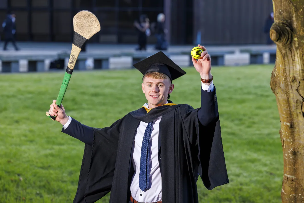 A photo of a man in smart dress and dark graduation robes and cap, holding a hurley in one hand and a sliotar in the other